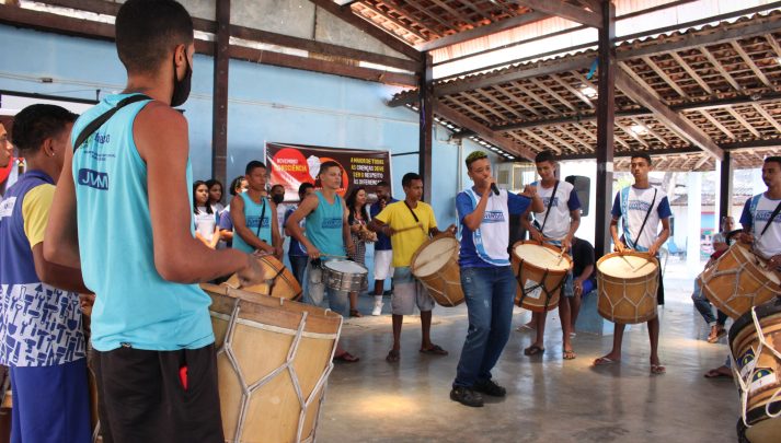Em um pátio, jovens do Centro da Juventude tocam alfaias em torno de um jovem trans negro, que canta segurando um microfone